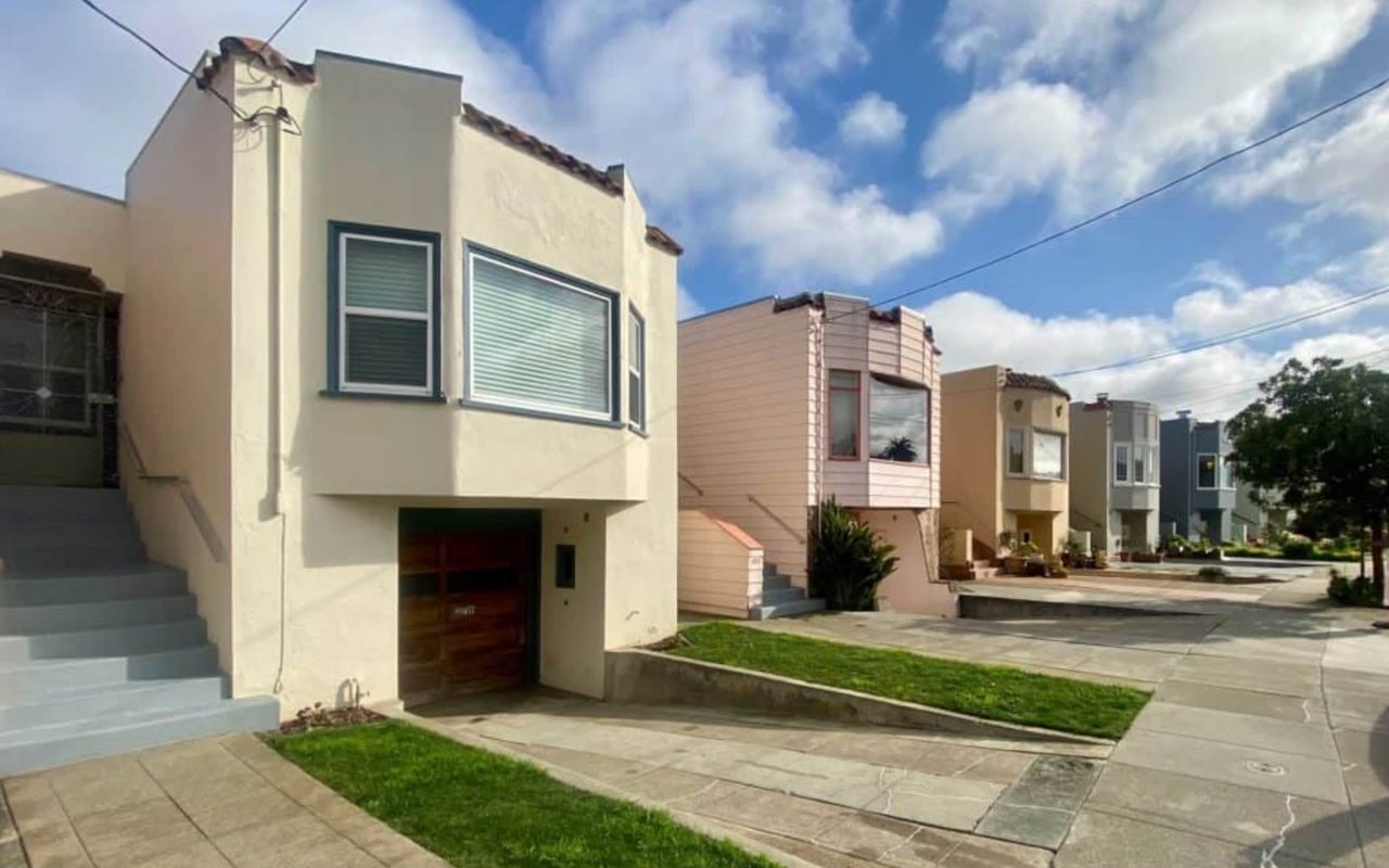 Cream two-story home with dark-trimmed windows beside a row of homes on a sunny street.