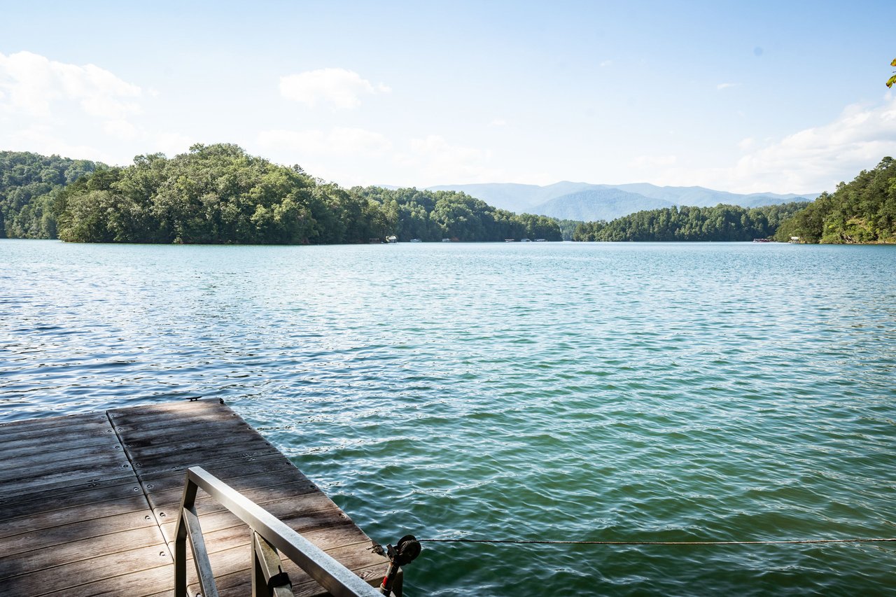 A wooden dock extends into a calm, greenish-blue lake surrounded by wooded hills under a clear sky.