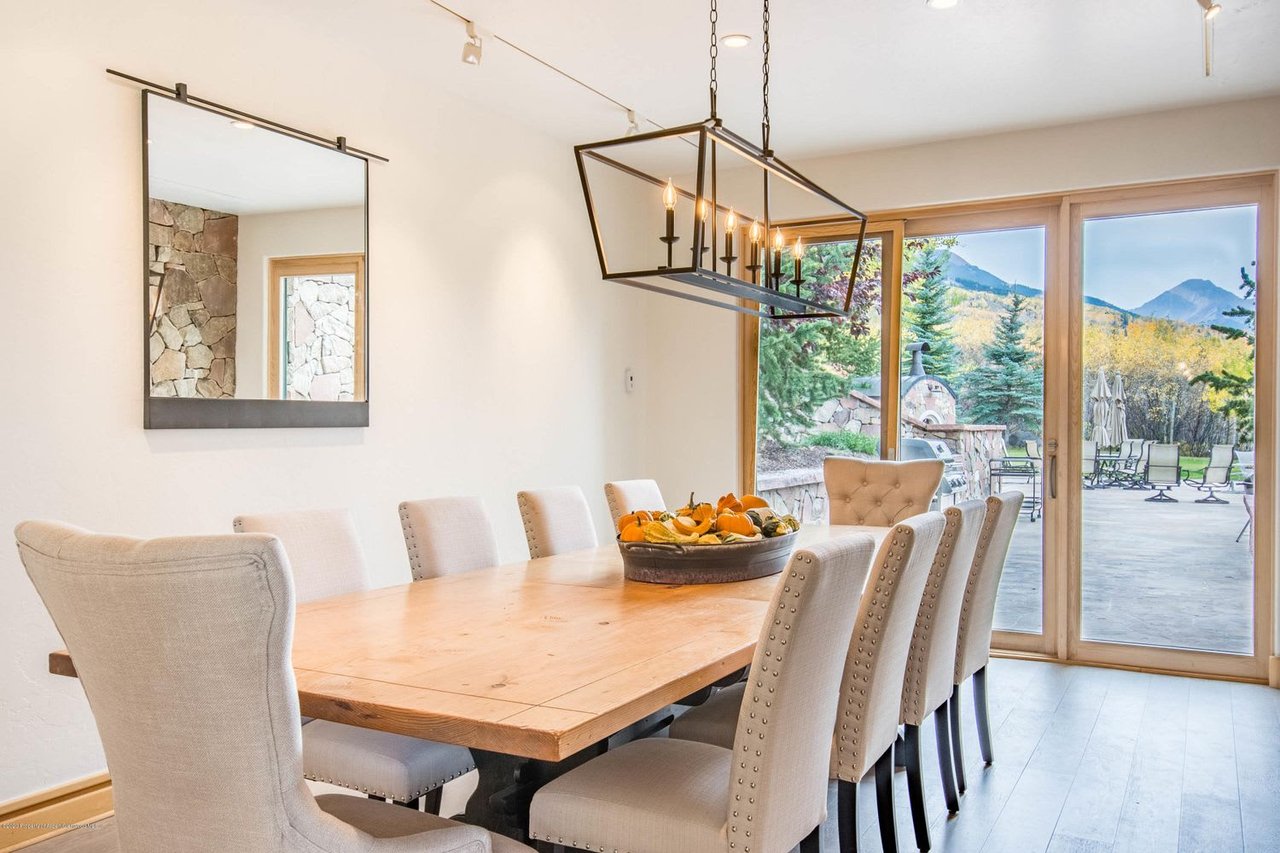 Dining room with a long wood table, upholstered chairs, and mountain views through glass doors.