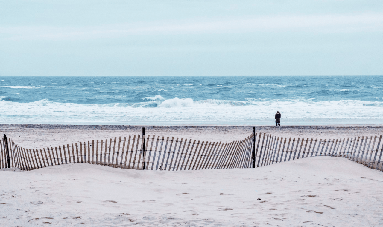 A person standing on the beach next to a fence.