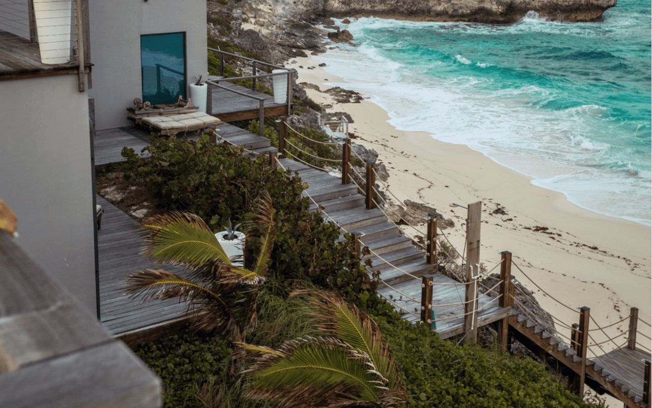  A narrow, weathered wooden staircase with a rope railing descends along a rocky cliff face, leading down to a secluded white sandy beach.