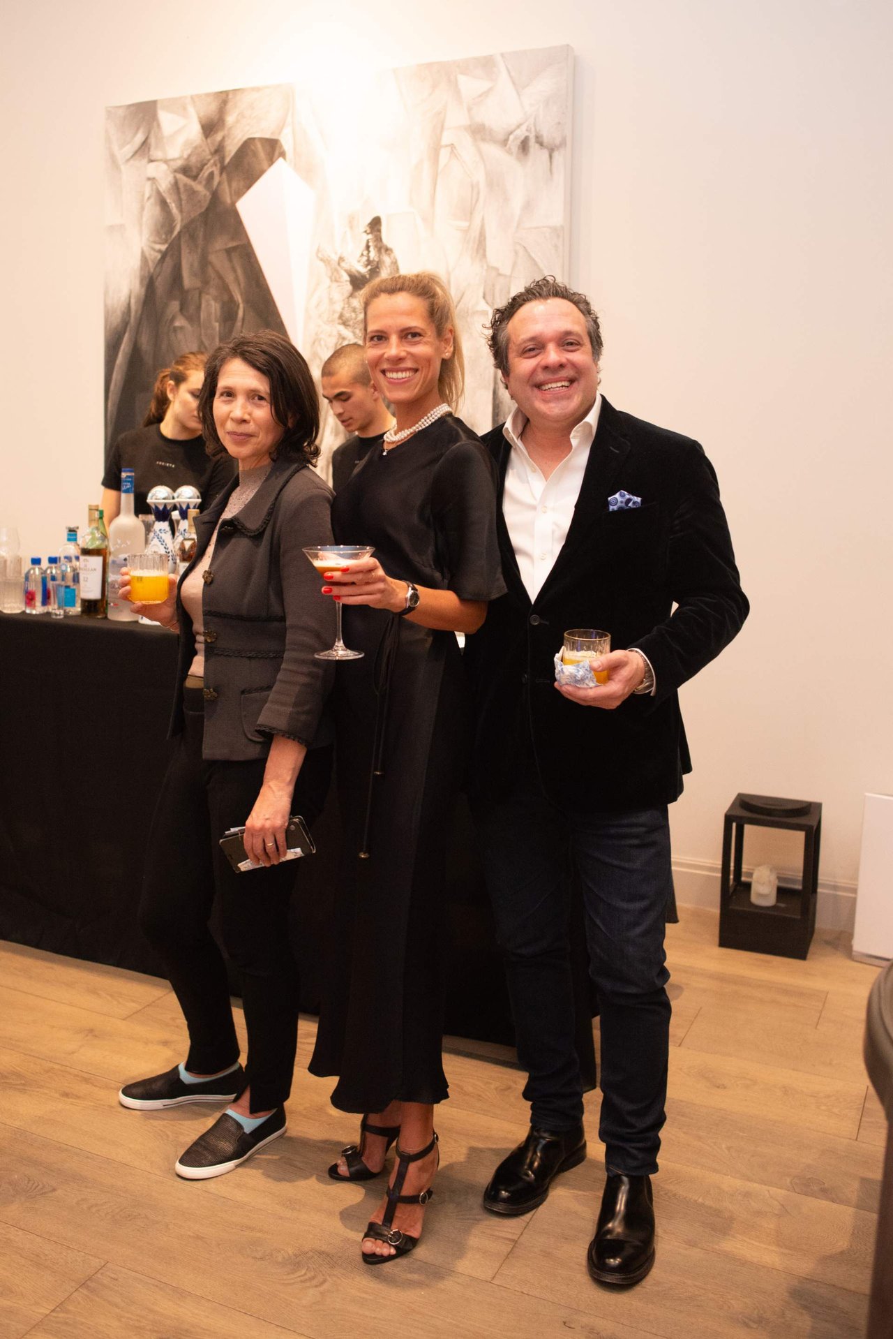 Three guests pose with drinks at an indoor reception, with a large abstract painting behind them.