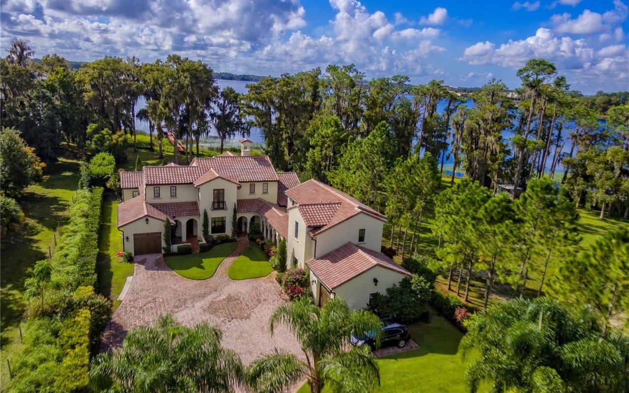 An aerial view of a large house with a two-car garage, a driveway, and a deck overlooking the water and surrounded by trees.