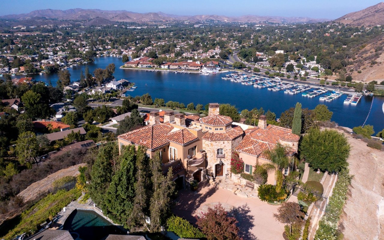 Aerial view of Mediterranean-style mansion on a hill with a pool, overlooking a lake with docked boats, surrounded by trees.