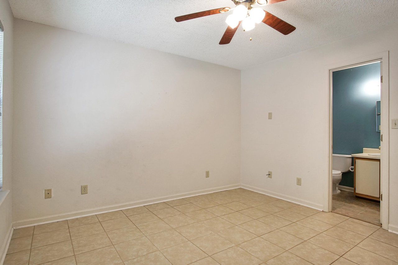 Photo of primary bedroom featuring light colored walls, a ceiling fan with light fixture, lightly tiled floors, a large window, and access to the primary bathroom  at 2709 Oak Park Court, Tallahassee, Florida 32308