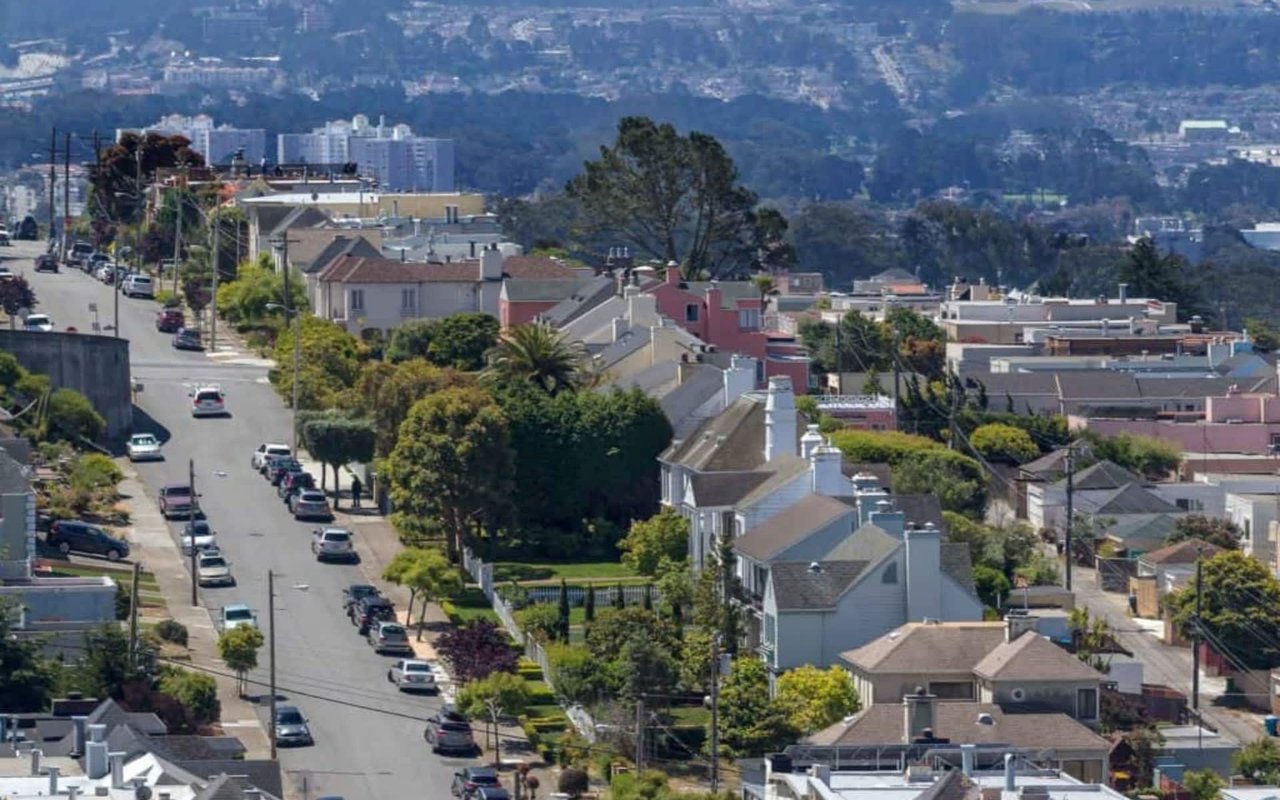 Residential street lined with houses and trees, with wooded hills in the distance.