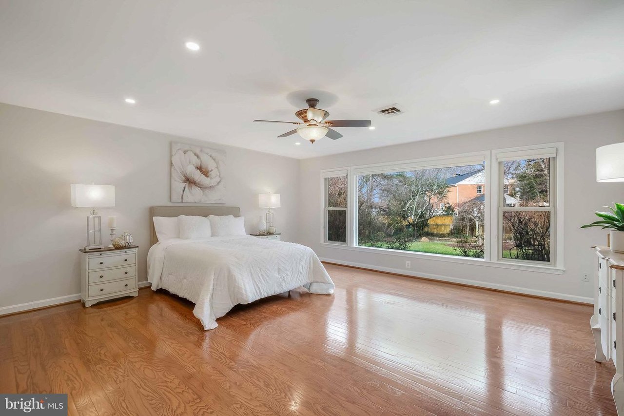 Staged bedroom with a white bed, hardwood floors, and large windows overlooking a yard.