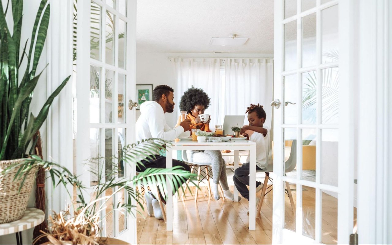 A view through a set of open white French doors into a bright, modern dining or living area where a family of three is seated at a white table. 