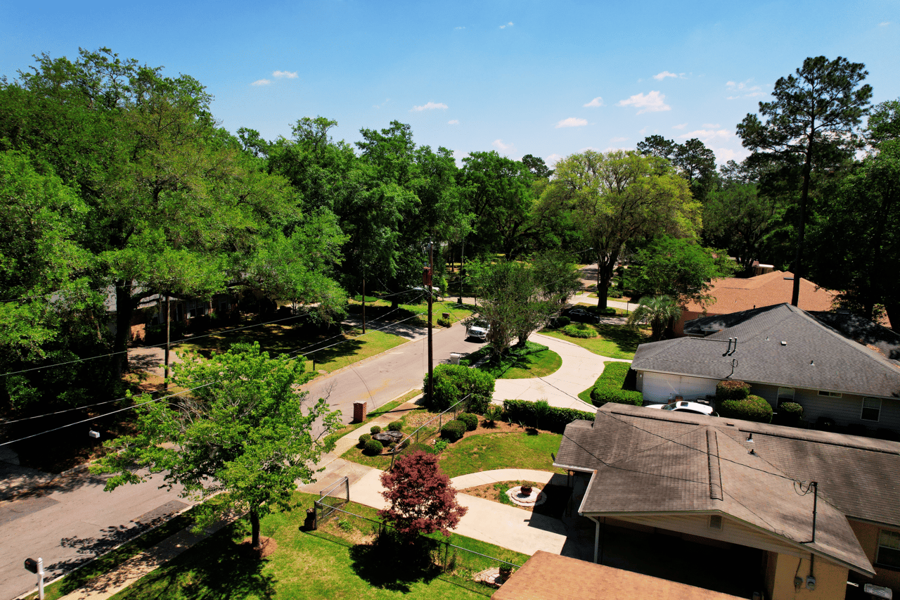 An aerial view of a residential neighborhood. The image shows multiple houses surrounded by trees and greenery, giving a suburban feel.