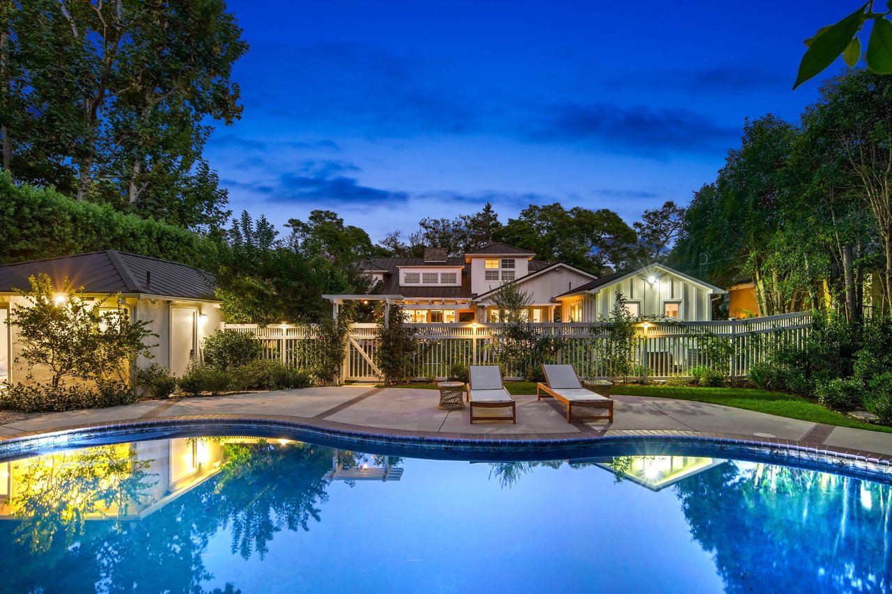  An illuminated swimming pool with a curved edge dominates the foreground of a spacious backyard