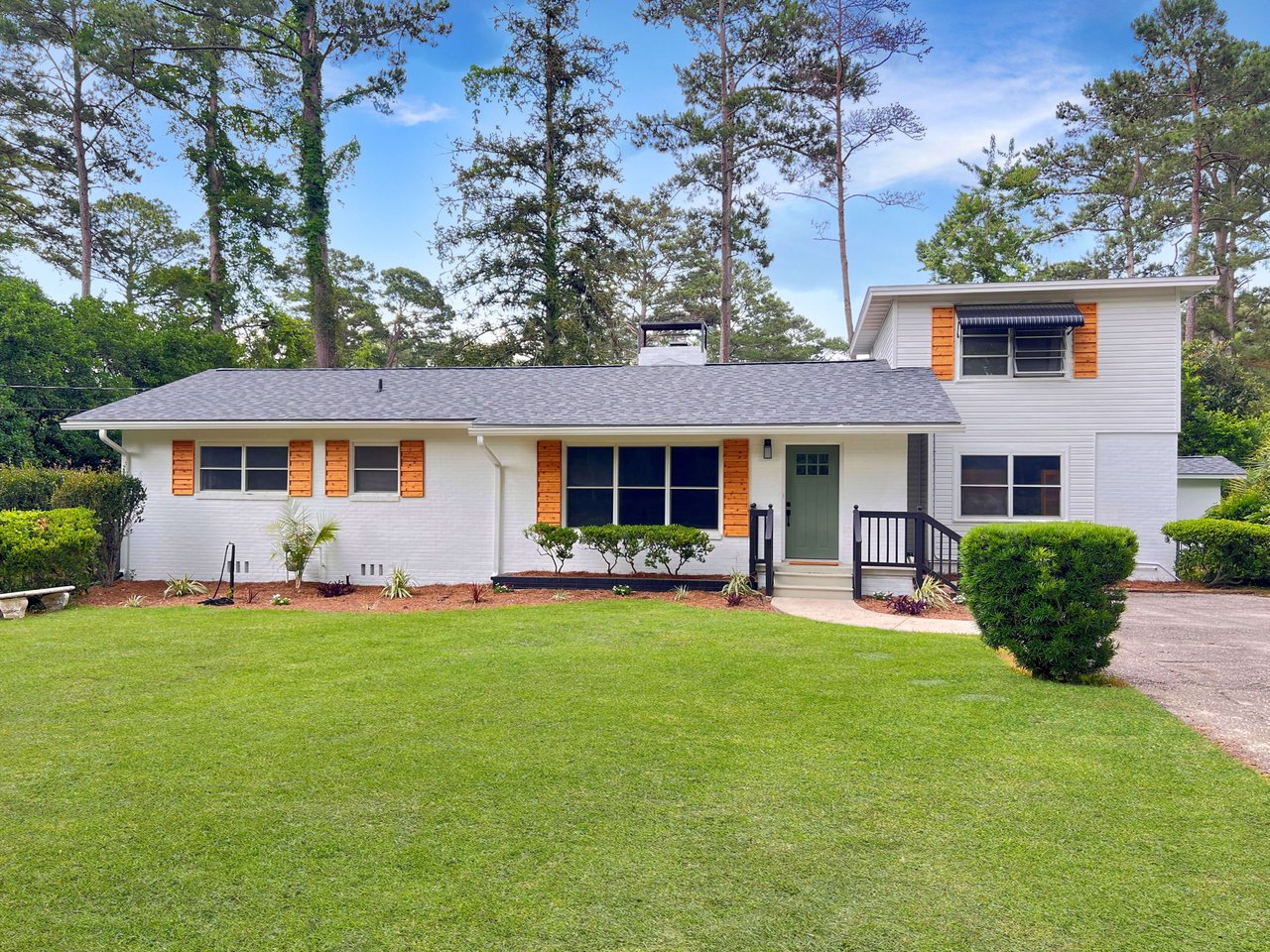 A well-maintained two-story house with a combination of brick and wood siding. The roof appears to be in good condition with dark shingles. The yard is well-manicured with green grass and neatly arranged plants and shrubs. There are a few young plants and a palm tree near the house.