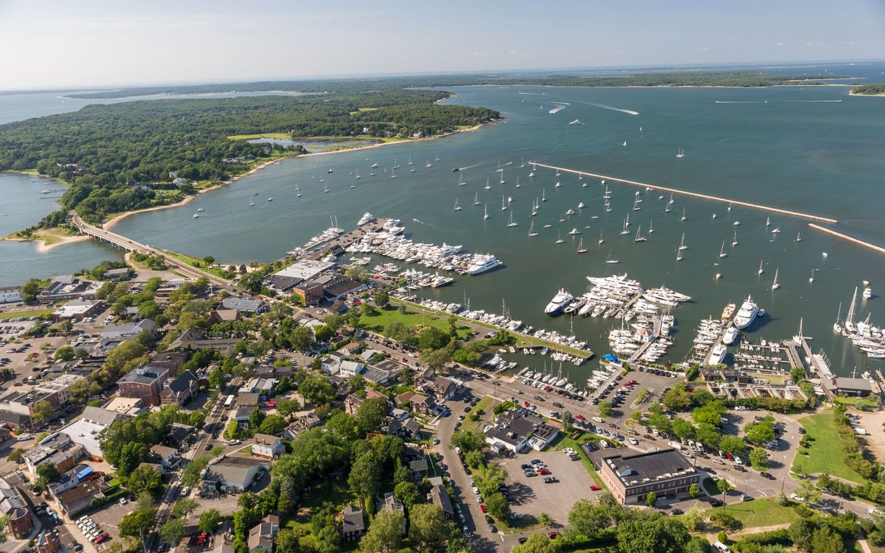 Aerial view of Sag Harbor, New York, featuring boats, yachts, colorful houses and buildings, and tree-surrounded areas.