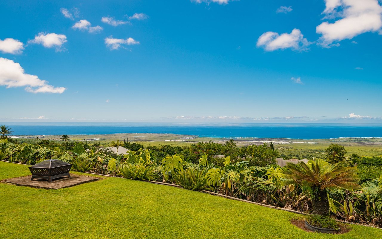 A lush green field with a stone fire pit and a view of the ocean