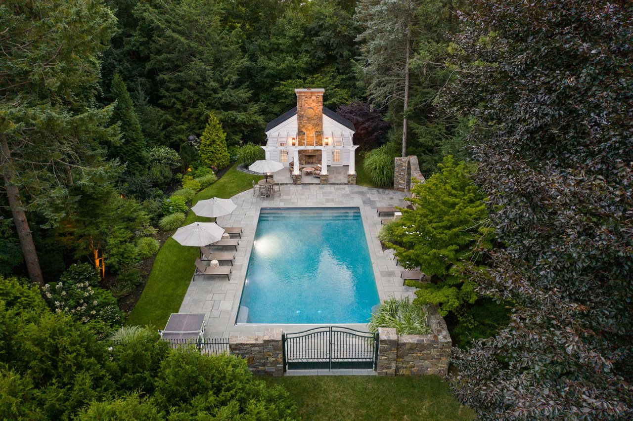  An aerial view of a luxurious backyard featuring a rectangular swimming pool with light blue water, surrounded by a gray stone patio.