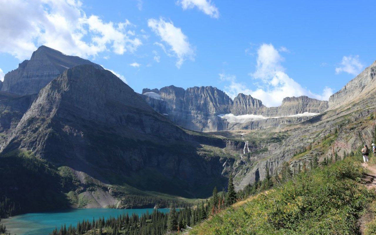A hiker on a trail that is narrow and winds through rocky mountains with evergreen trees.