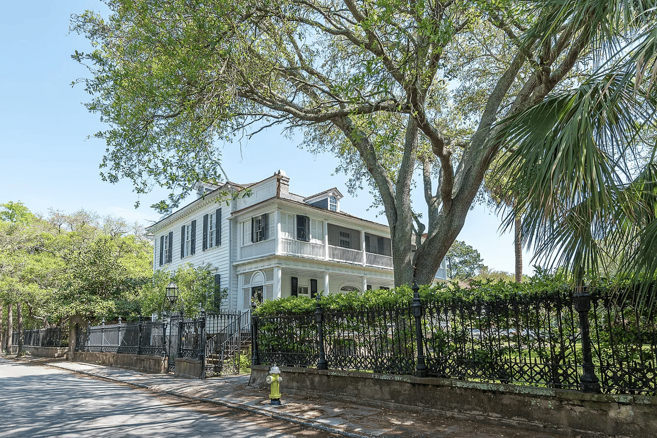 White two-story house with a wraparound balcony behind an iron fence and mature trees.