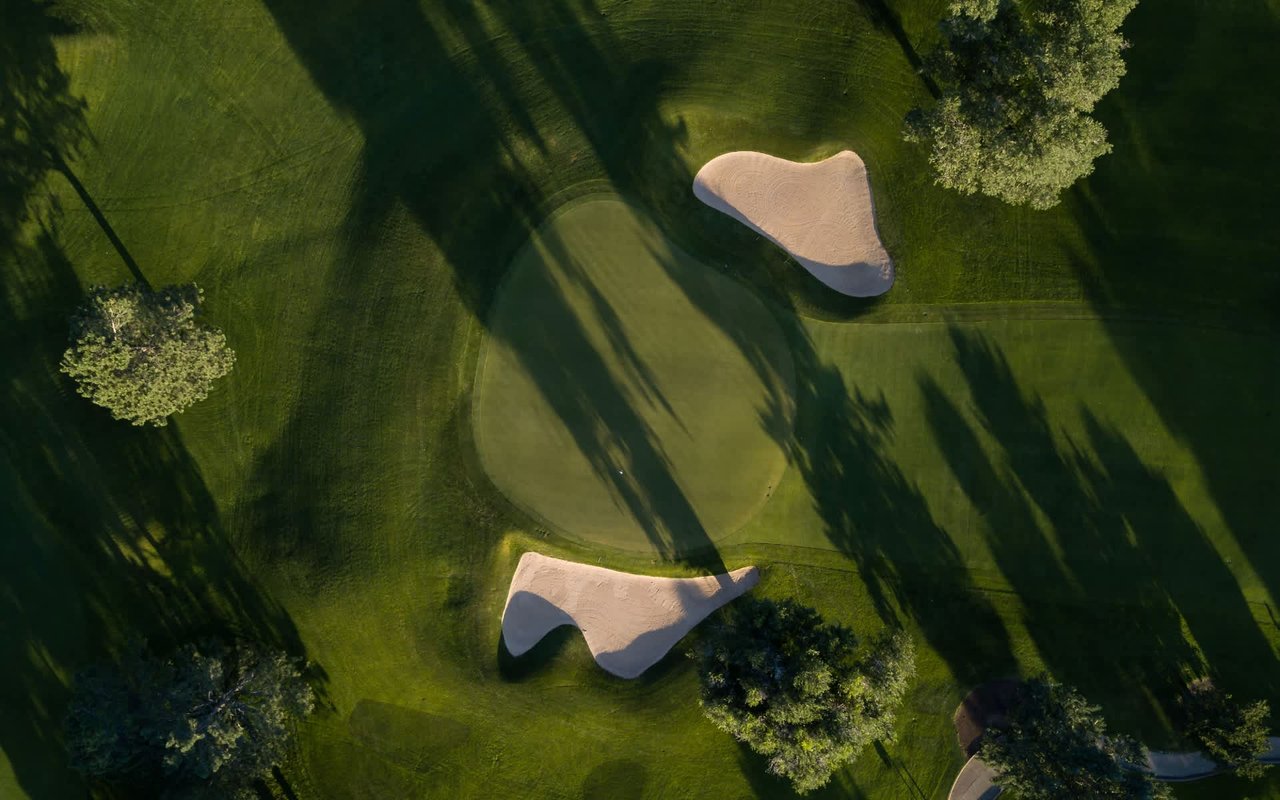  An aerial view of a golf course features a dark green putting green surrounded by lighter green grass.