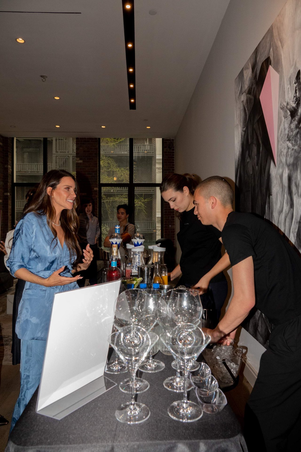 Guests gather at a tasting counter, with a woman in blue speaking to two drink servers.