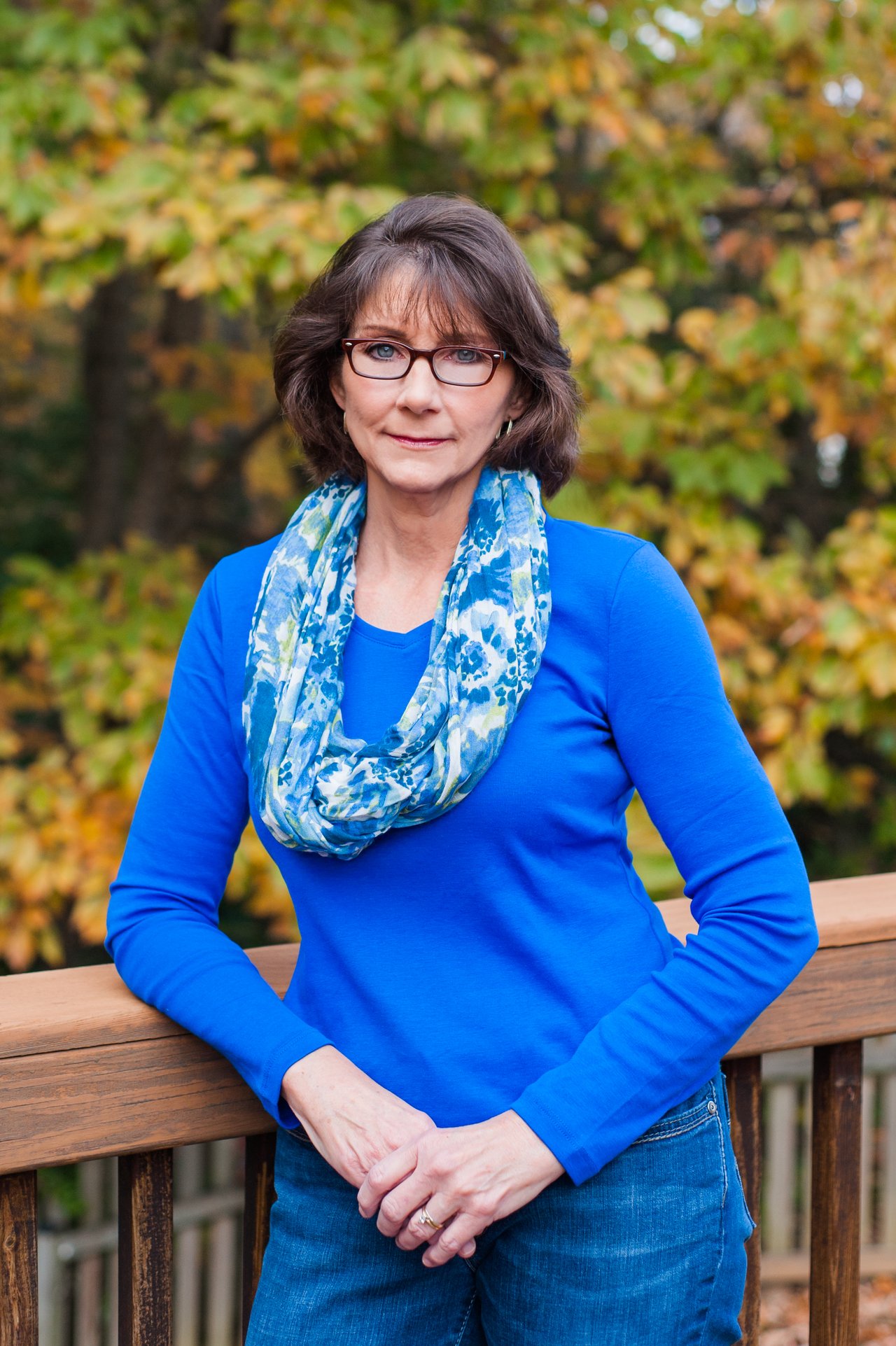 Dawn Ferriss in a blue top and patterned scarf, standing outdoors before autumn foliage.