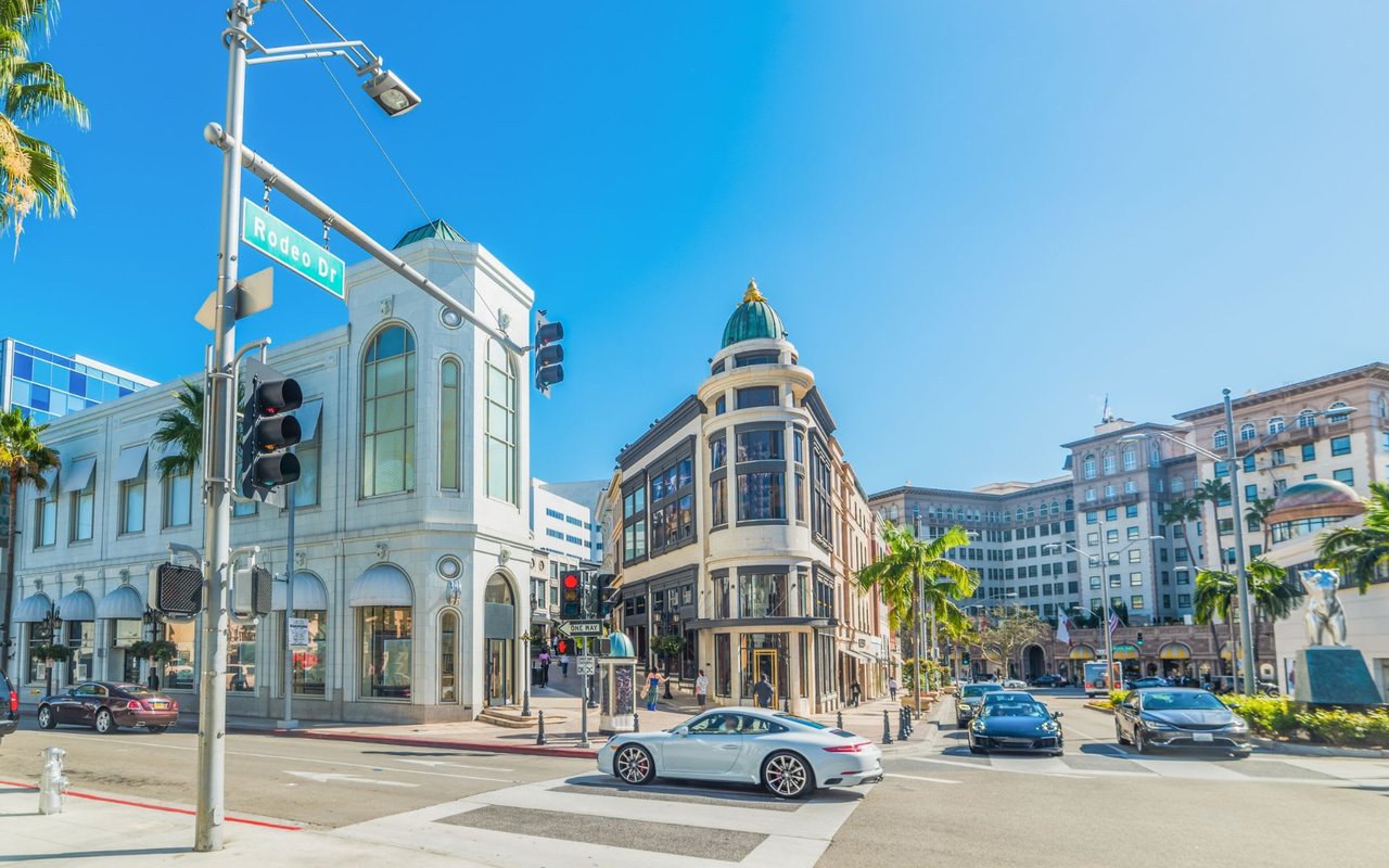 Rodeo Drive in Beverly Hills features luxury shops, palm trees, white stone buildings, and few visible cars and people.