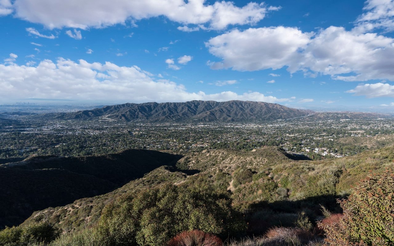 A panoramic view of a city from a mountaintop. The city is surrounded by lush green mountains and forests.