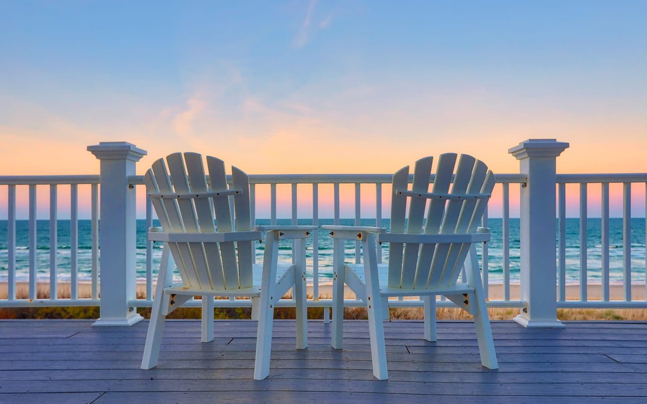 Two white lawn chairs sitting on a wooden deck overlooking the ocean.