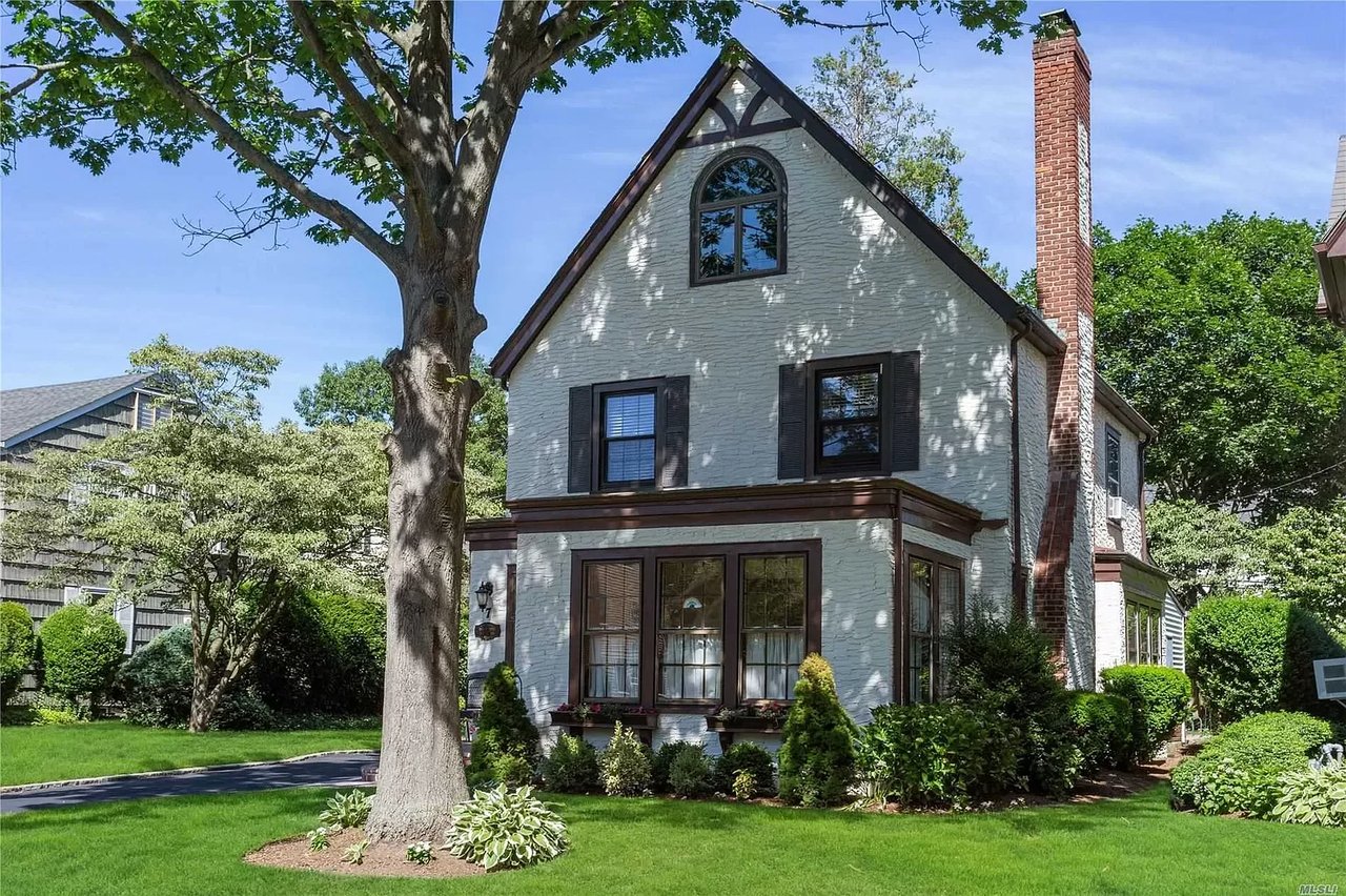 White stone house with a steep gable and brick chimney behind a large shade tree.