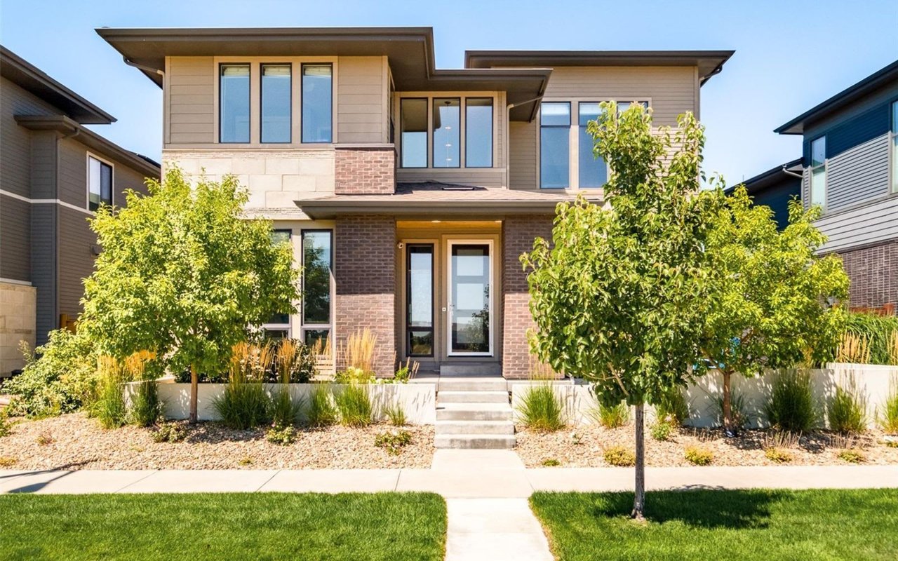  A large, modern suburban house with light brown and grey siding, dark brick accents, and a contrasting brown roof 