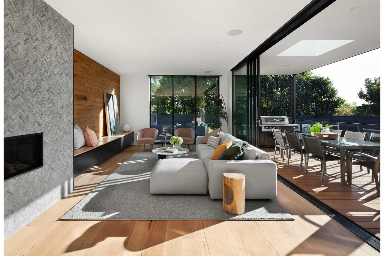 Modern living room with an open-concept layout, featuring large sliding glass doors that lead to a patio. The space includes a neutral color palette, light gray sectional sofa, textured gray rug, warm wood accents, stone fireplace column, and outdoor dining area with a grill.