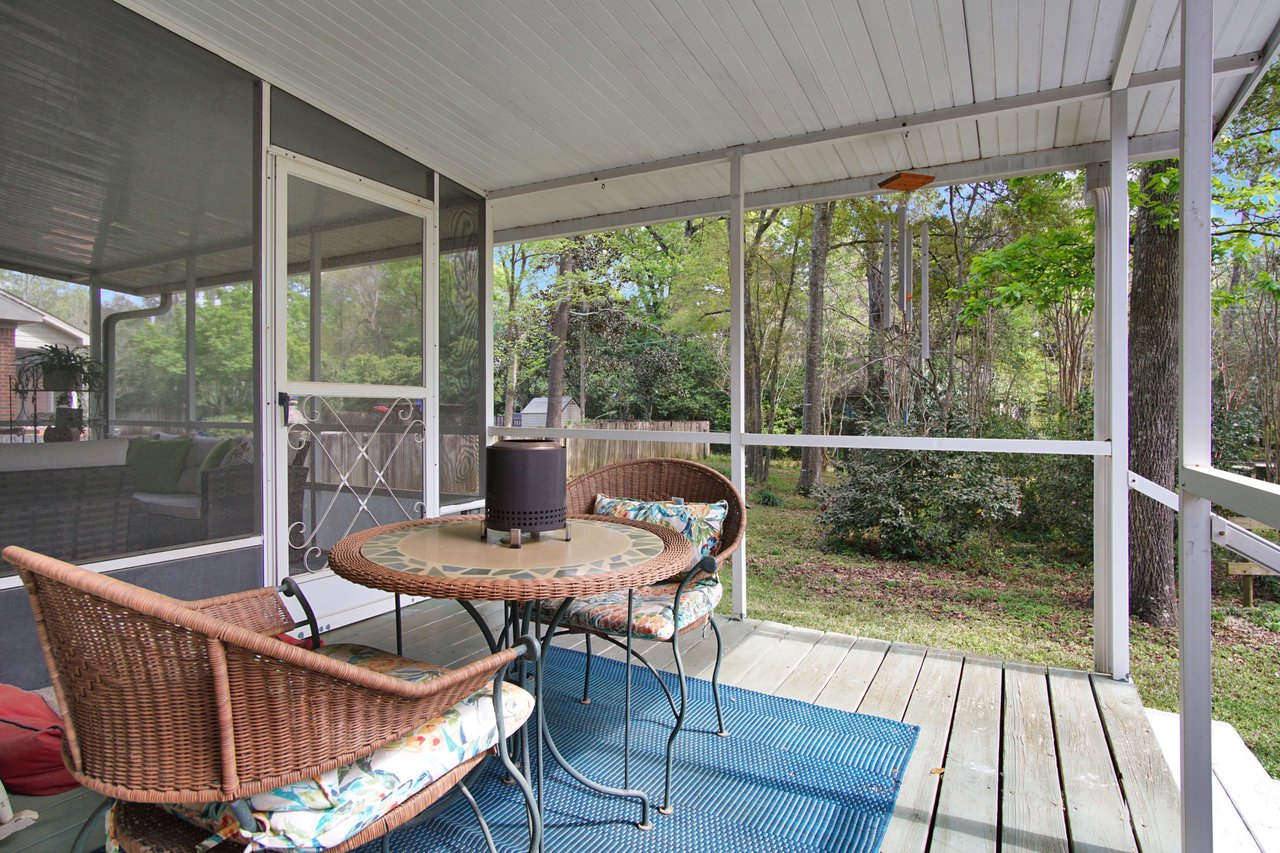 A screened porch or sunroom with wicker furniture and a table. The area is cozy and provides a nice view of the surrounding greenery.