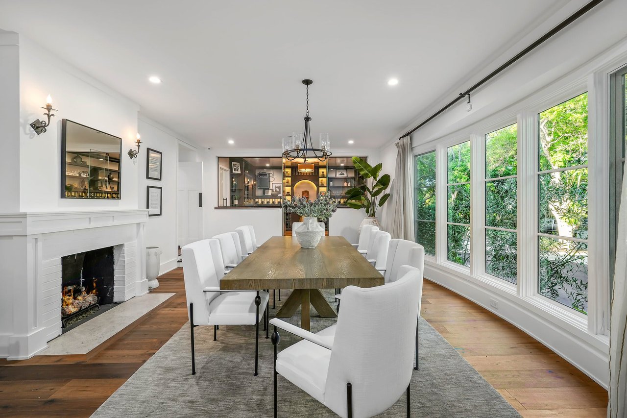 A spacious and elegantly decorated dining room features a long rectangular wooden dining table with a thick base, surrounded by eight white upholstered chairs.