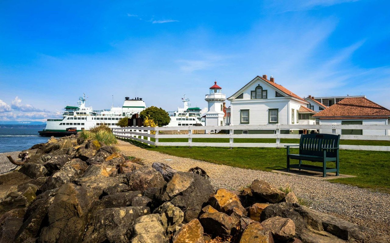 Mukilteo lighthouse beside the rocky shore, with ferries in the background and a bench on the lawn.