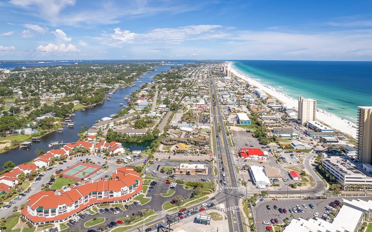 An aerial view of Panama City Beach, Florida, showing a long stretch of white sand beach, turquoise water, and a bustling coastal town. 