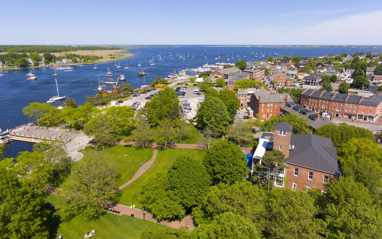 A wide blue body of water, possibly a bay or river, is filled with numerous boats and yachts, including what appears to be a replica tall ship docked near the shore. 