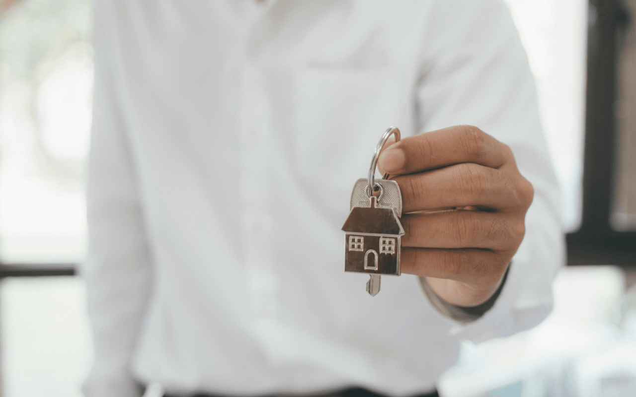  A person wearing a white long-sleeved shirt is holding out a set of keys with a house-shaped keychain attached. 