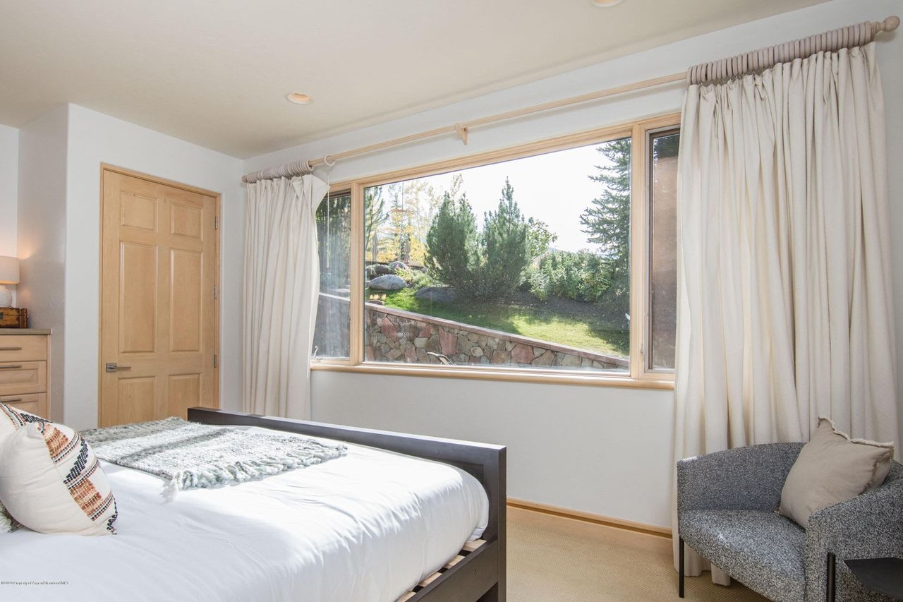 Furnished bedroom with a large window overlooking a landscaped yard and stone retaining wall.