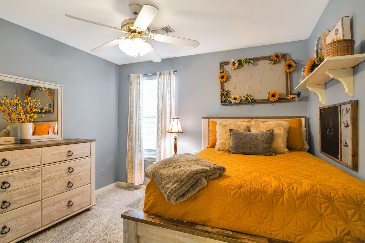 A bedroom with a yellow bedspread, blue walls, and wooden furniture. The room is decorated with shelves, artwork, and has a ceiling fan.