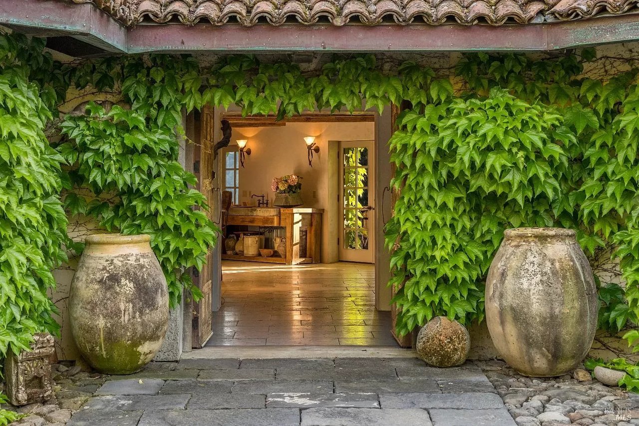 Entrance of a rustic home with ivy-covered walls and large ceramic pots, leading to a warmly lit interior.