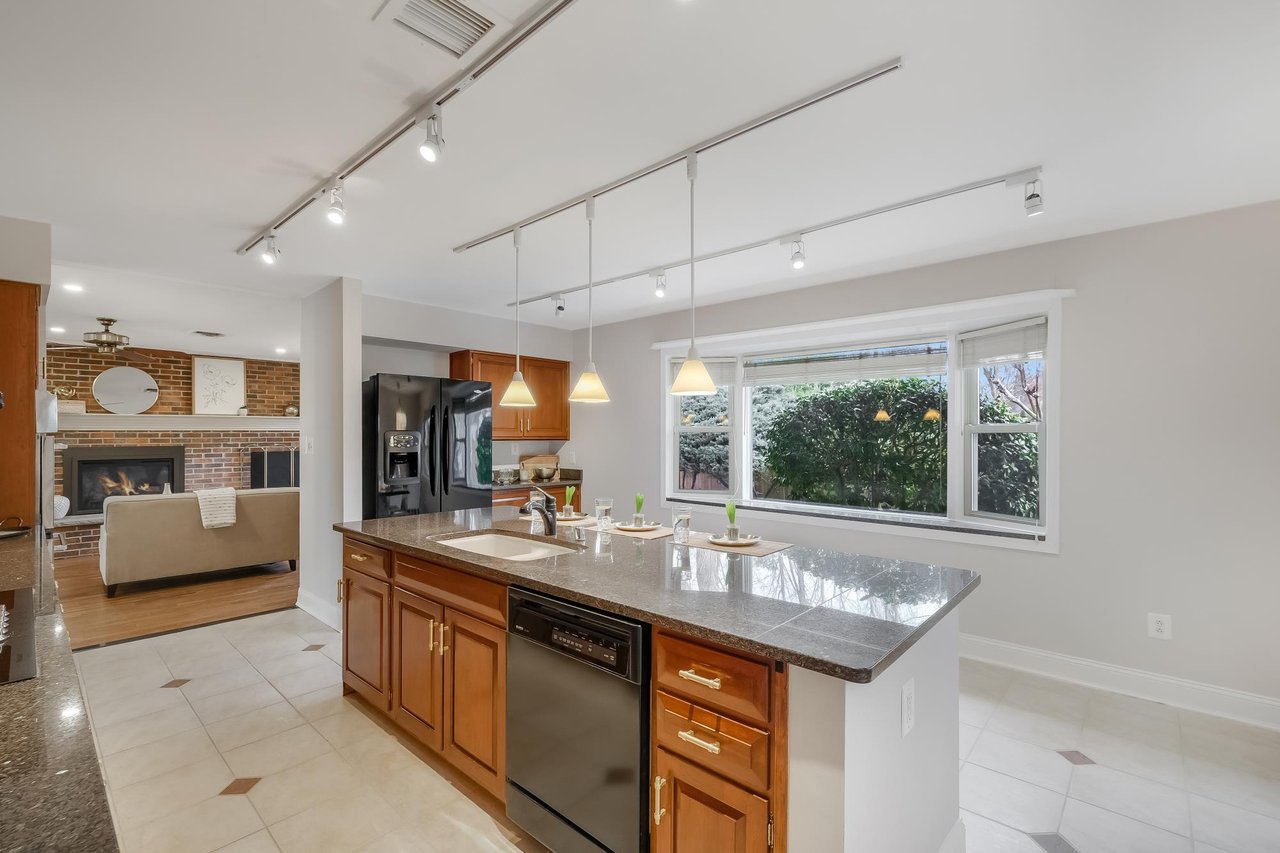 Kitchen with a granite island, wood cabinetry, pendant lights, and an adjoining living room with a brick fireplace.