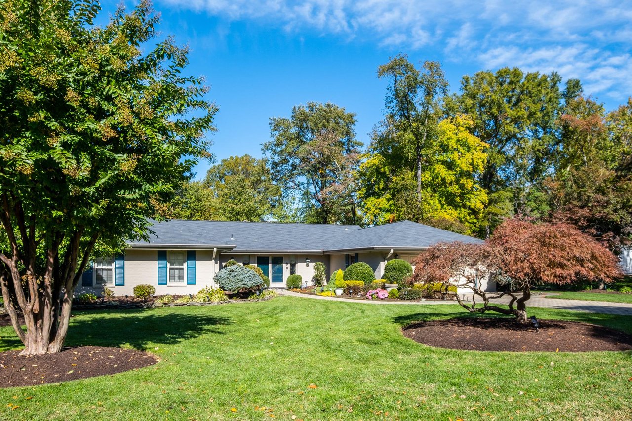 Single-story home with a gray roof, landscaped front yard, mature trees, and curved driveway.