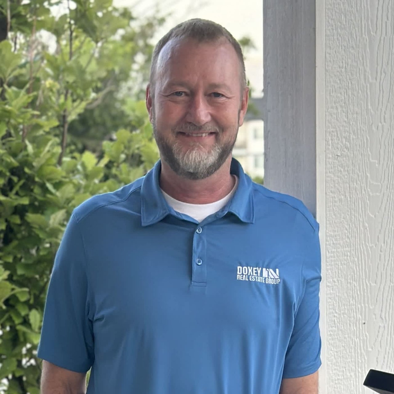 A man with a beard, wearing a light blue polo shirt with the logo "Doxey Real Estate Group" on the chest. He is standing outside, smiling, with his hands by his side. There is greenery in the background, and he is standing next to a white column.