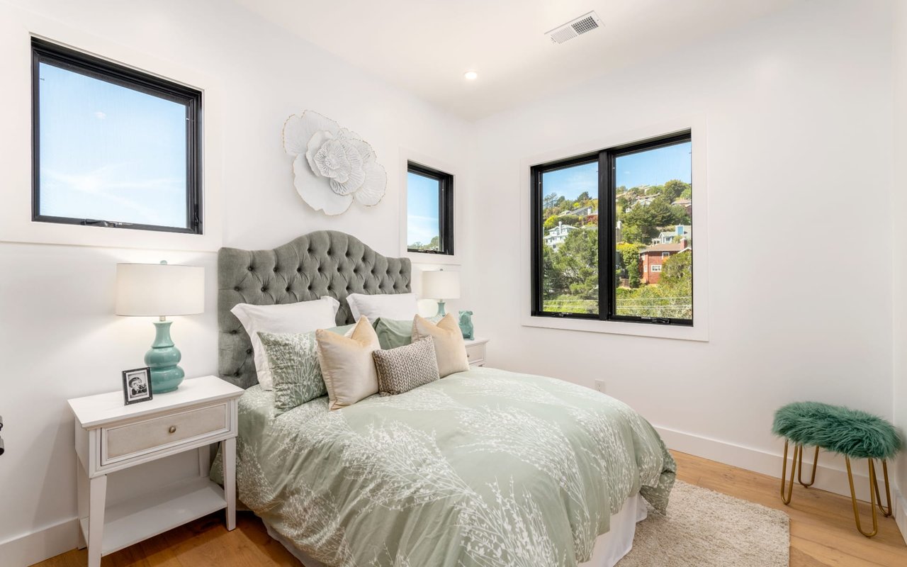 Staged bedroom with pale green bedding and windows overlooking a leafy hillside neighborhood.