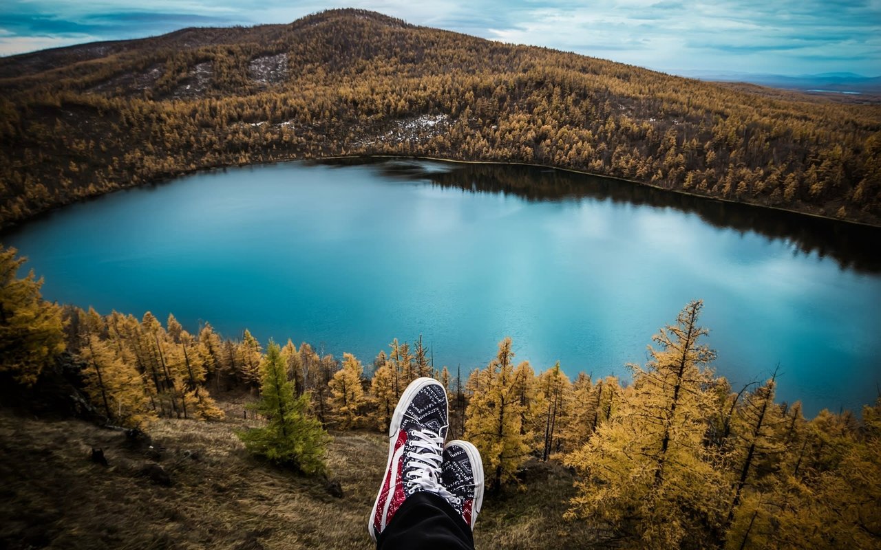  A pair of feet, clad in black pants and white and red patterned sneakers with white laces, are propped up in the foreground, overlooking a stunning circular blue lake. 