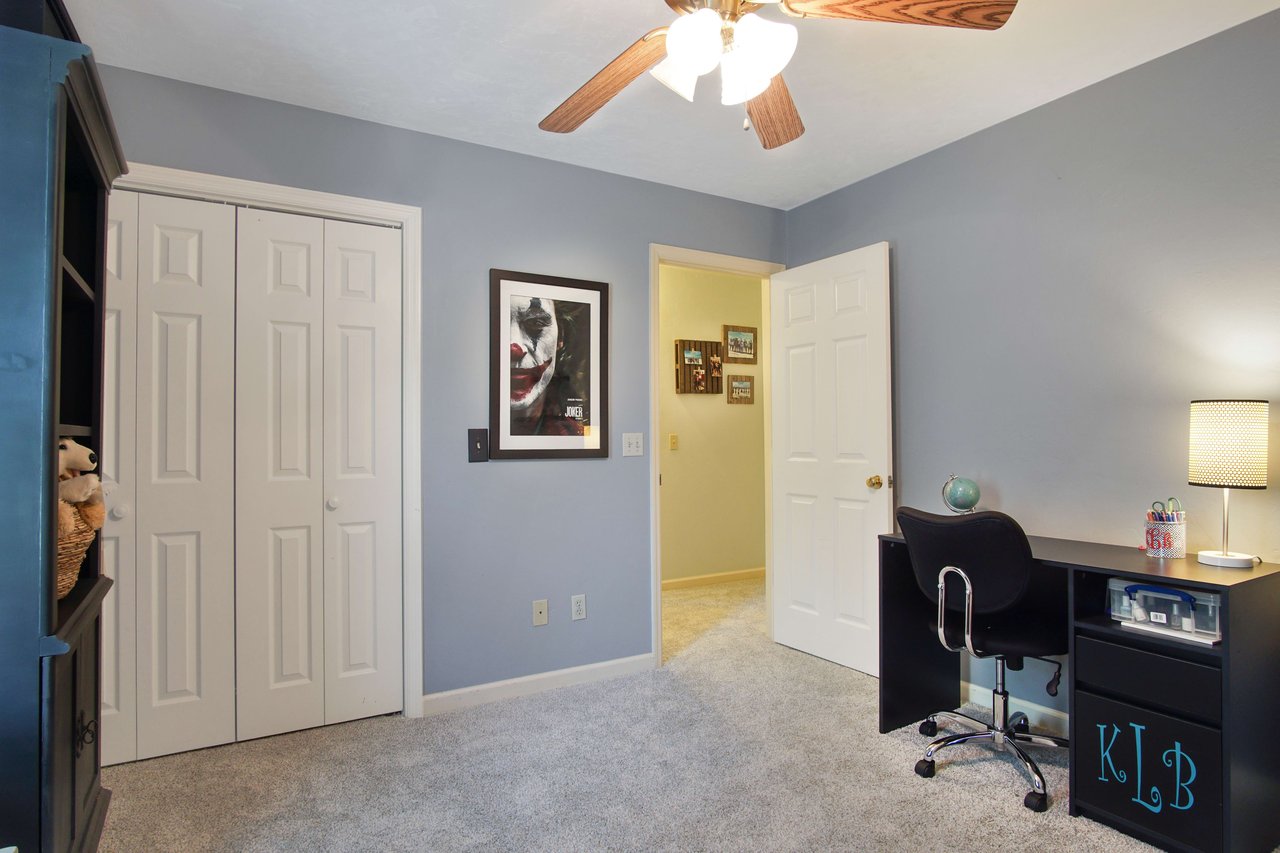 An interior shot of a bedroom with blue walls, carpeted flooring, and a ceiling fan. The room includes a desk and chair, and there is a closet with white doors.