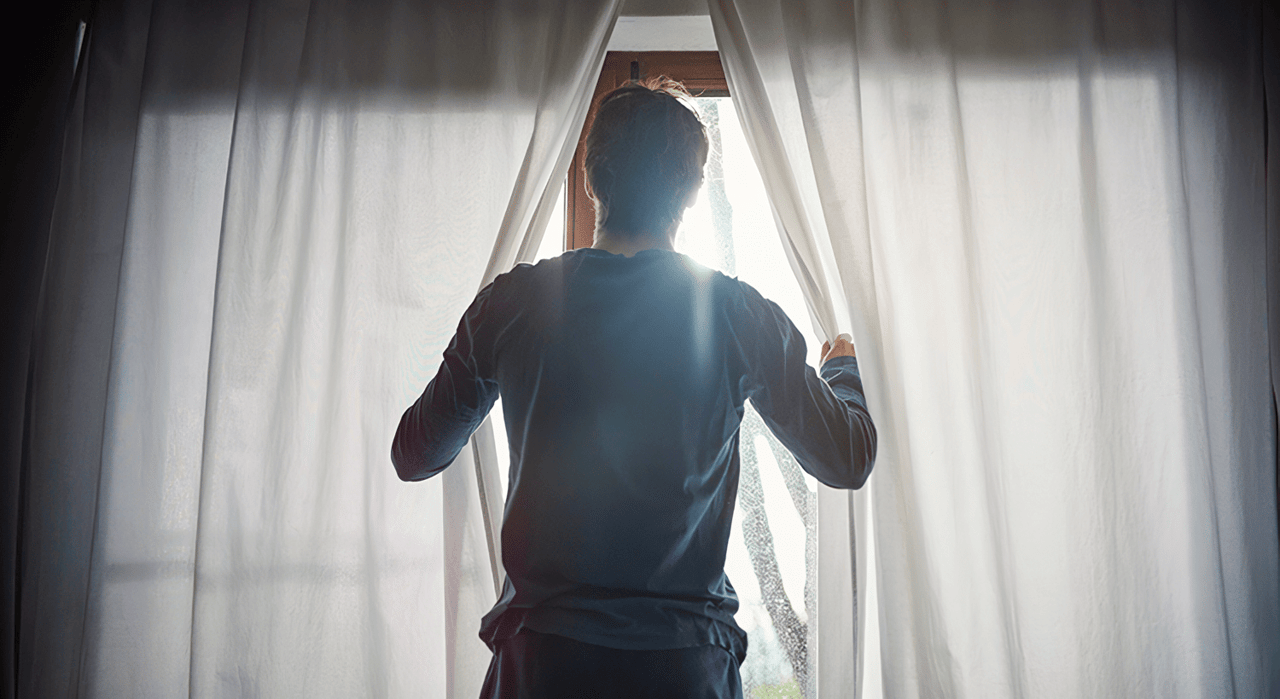 A guy in blue pajamas parting white curtains looking out the window