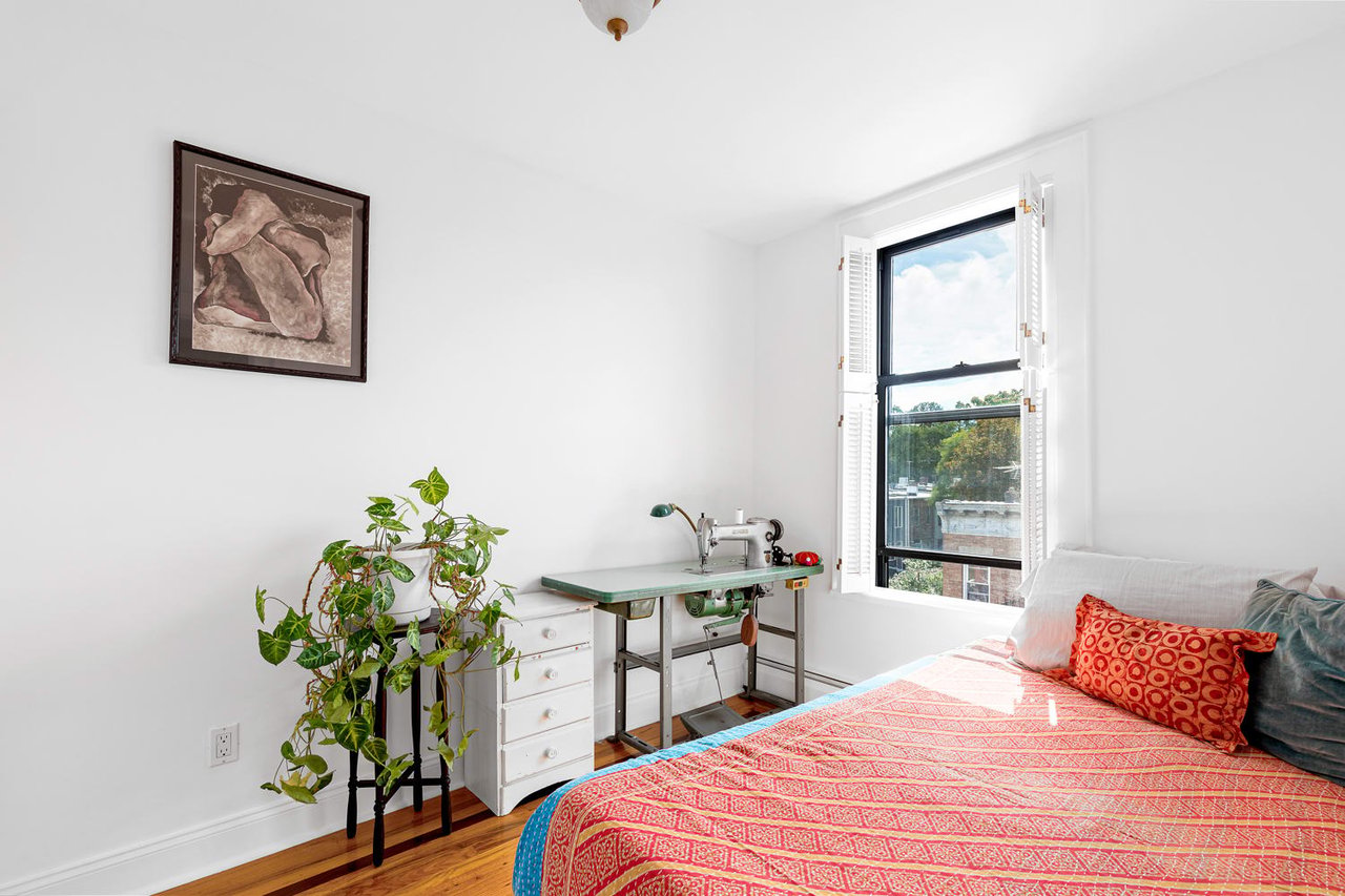 Bedroom with a red patterned bedspread, sewing machine table, and a large shuttered window.
