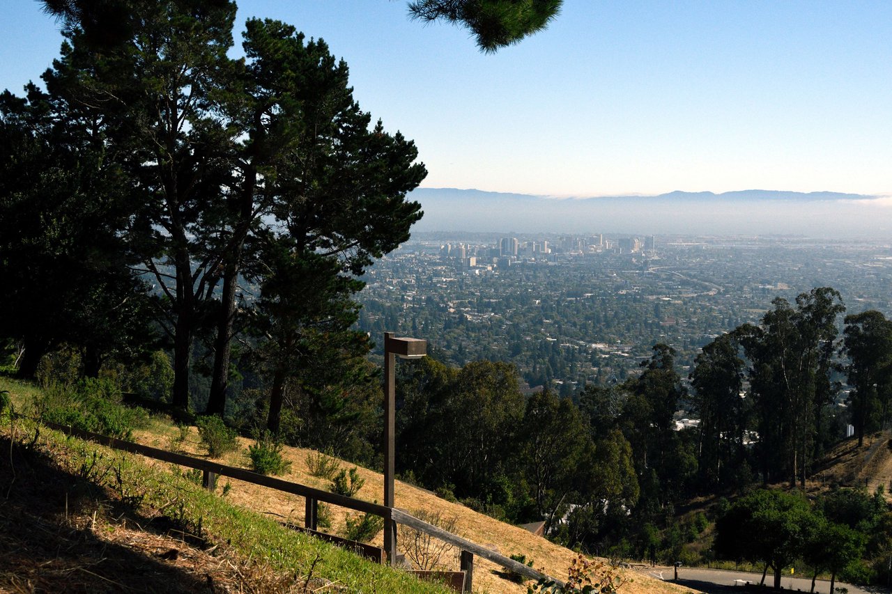 Panoramic view of a city from the top of a hill, with tall buildings, mountains, and a blue sky.