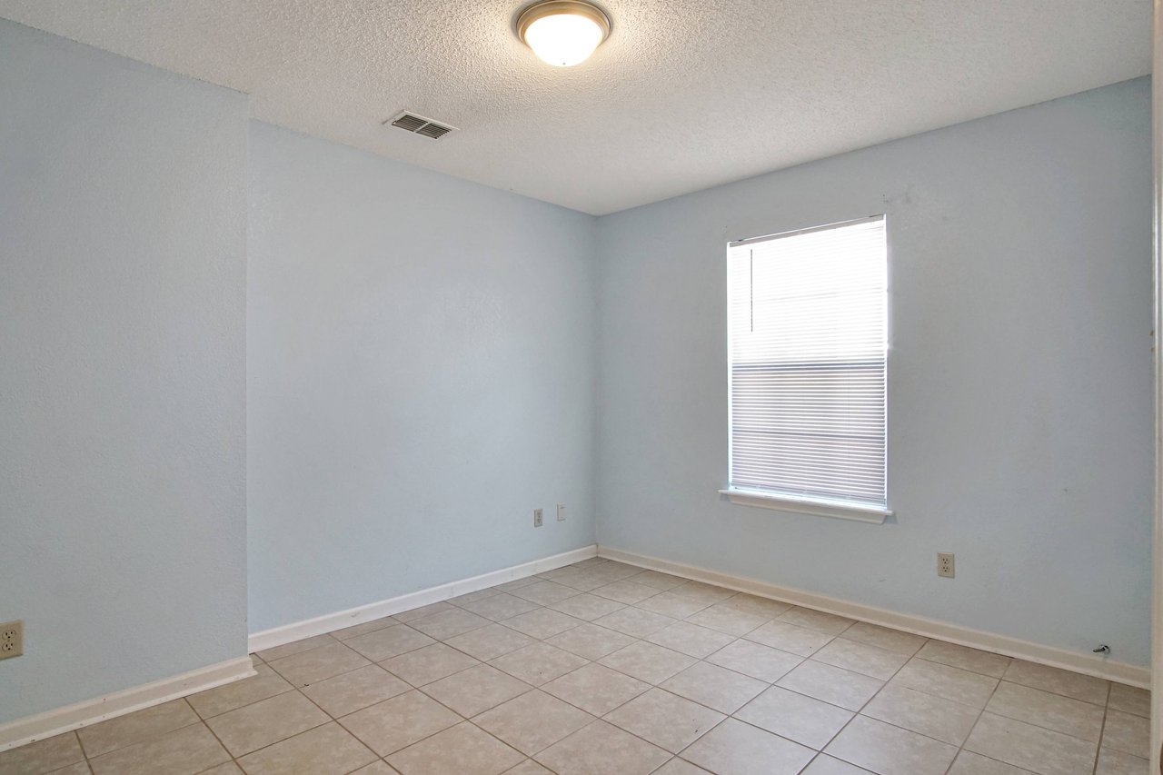 Photo of the second bedroom featuring light blue walls, a white ceiling, lightly colored tile, a window with white blinds, and a simple light fixture  at 2709 Oak Park Court, Tallahassee, Florida 32308