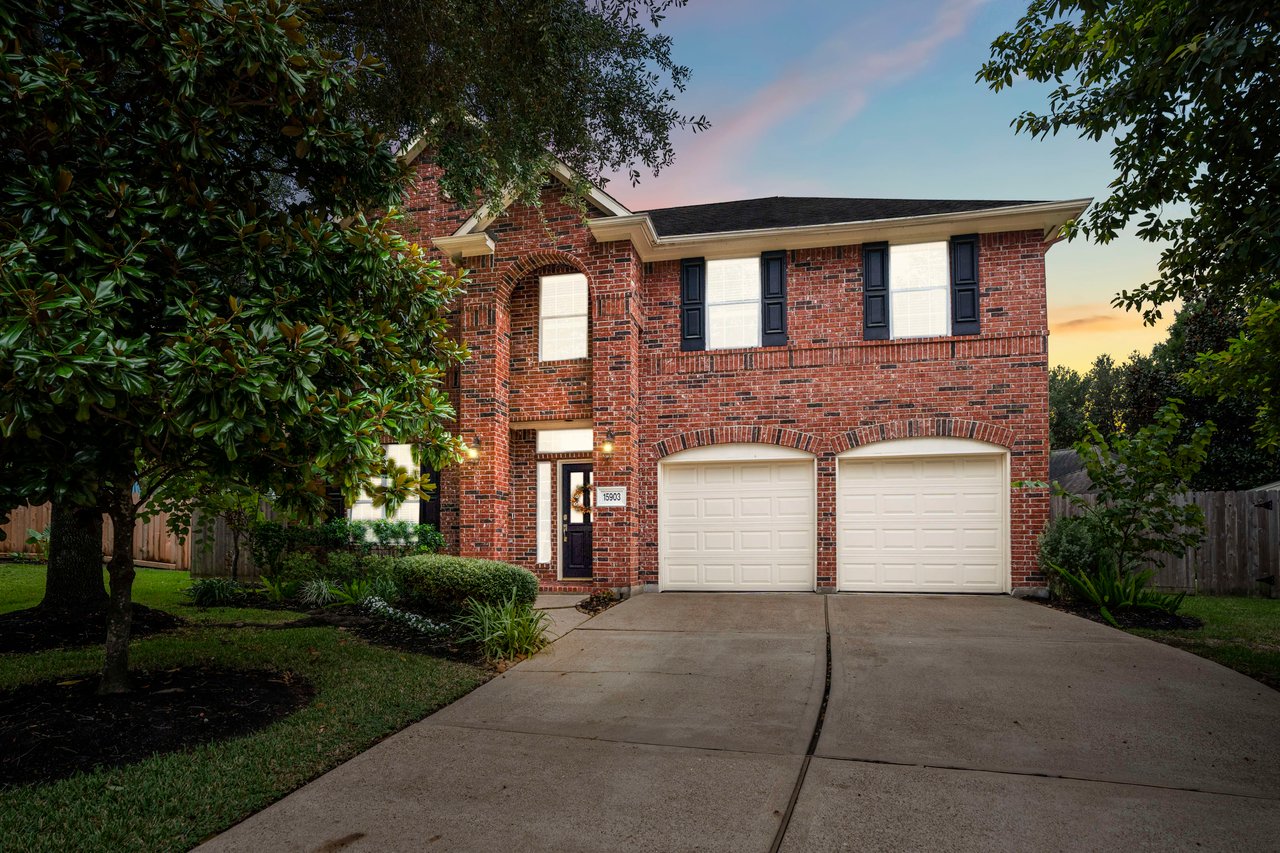 Two-story red brick house with a double garage, broad driveway, and mature trees at dusk.
