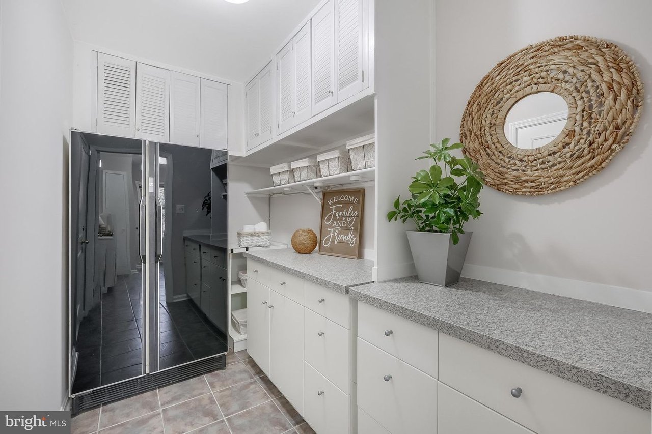 Kitchen pantry with white cabinets, gray counters, stainless-steel refrigerator, and woven round mirror.